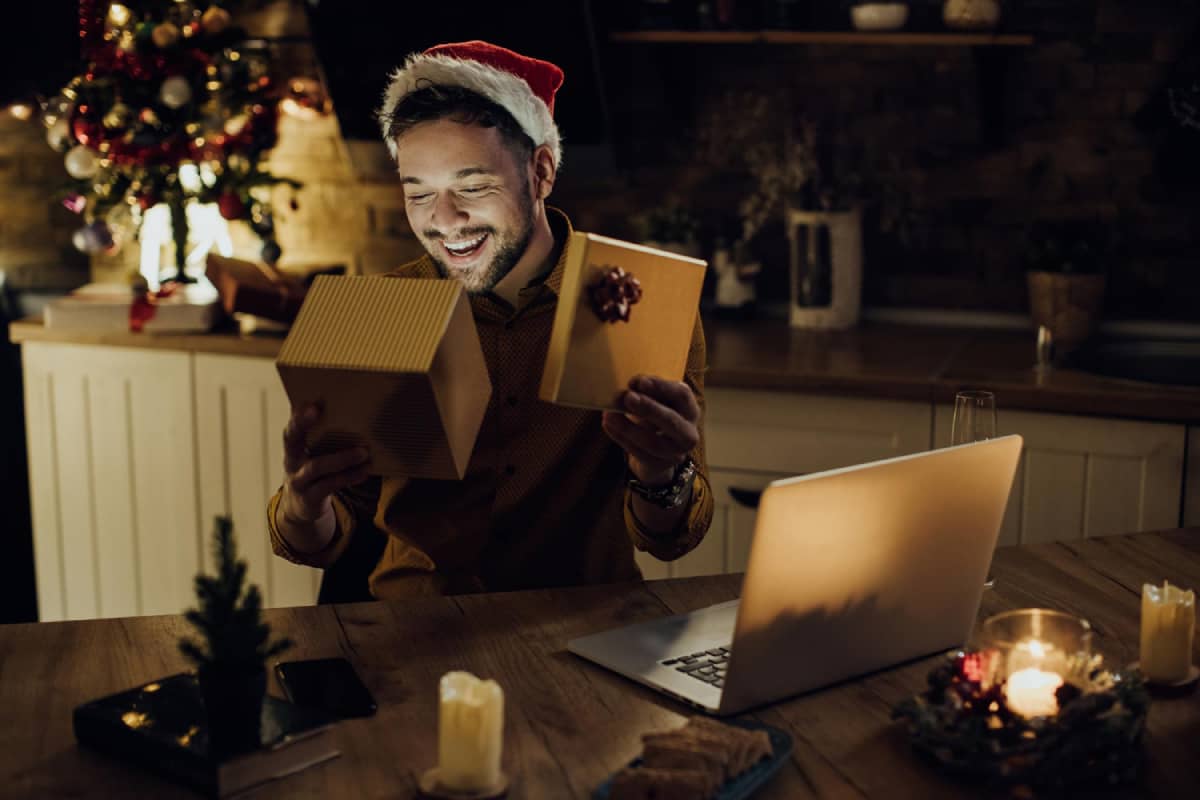 Cheerful man in a christmas hat opening a present behind his laptop