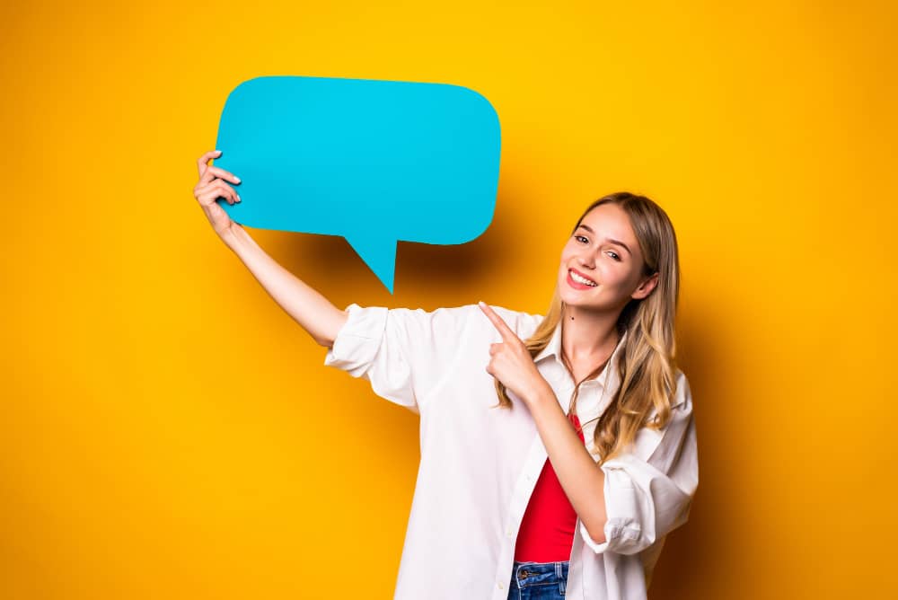 Happy young woman holding an empty blue speech bubble infront of isolated yellow wall