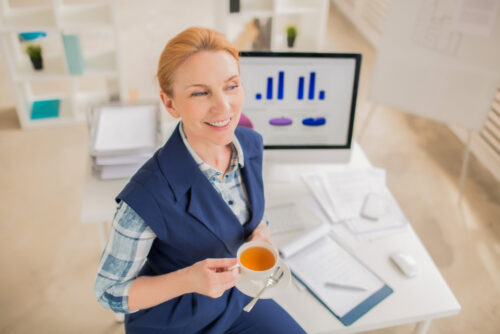 Email marketer drinking tea at their desk, happy at campaing results seen on the background