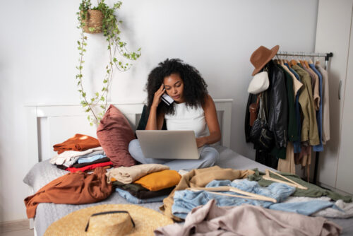 A woman doing online shopping on her bed full of clothes, looking fatigued