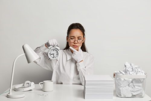 A woman holding a clock at a white cluttered desk in a fully white-colored room