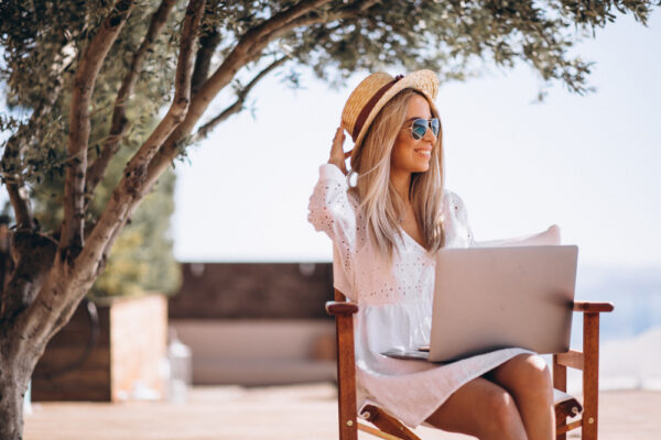 Woman in a white dress with sunglasses in the summertime, working on her computer and smiling