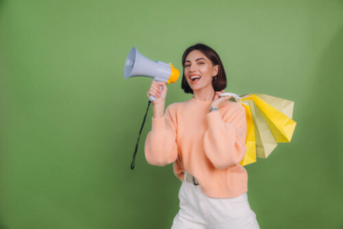Young woman in casual wear isolated on green olive color wall shouting in a megaphone, holding shopping bags, announcing discounts sale promotion